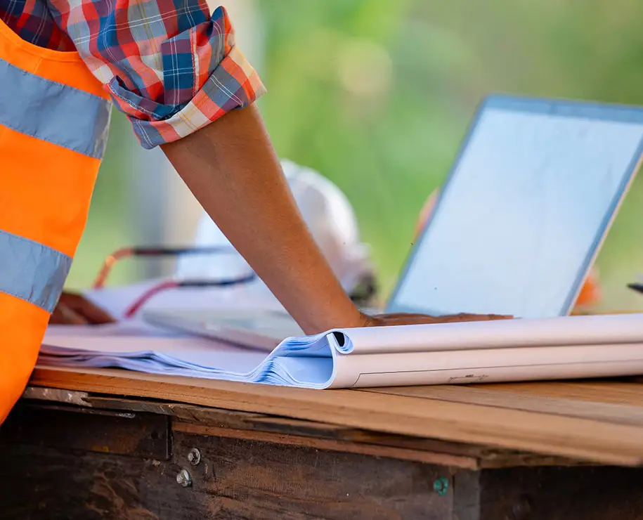 Ein Bauingenieur steht vor einem Tisch mit Bauplänen und Laptop