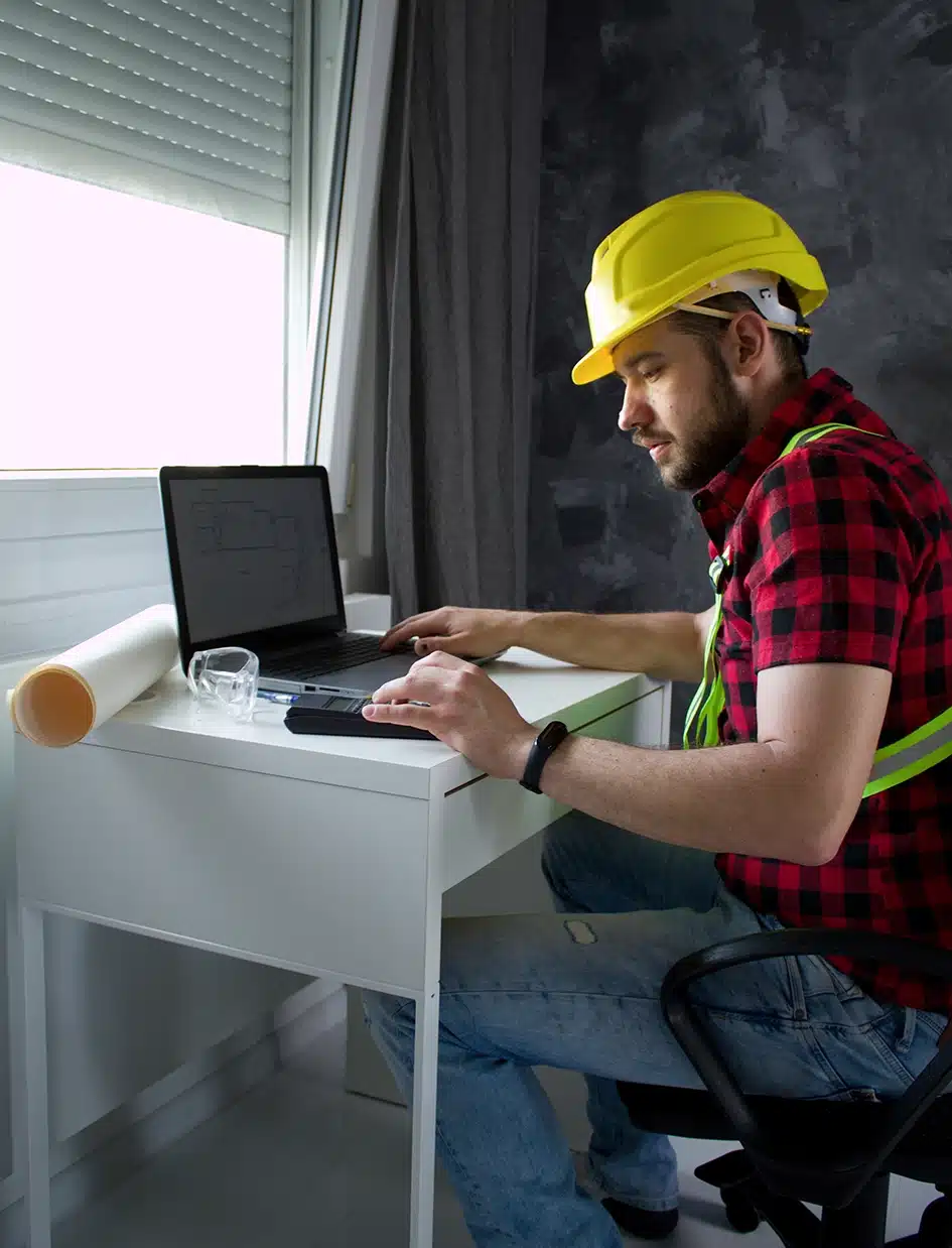 Bauarbeiter sitzt an seinem Schreibtisch am Computer, schaut sich Baupläne an und führt Rechnungen durch