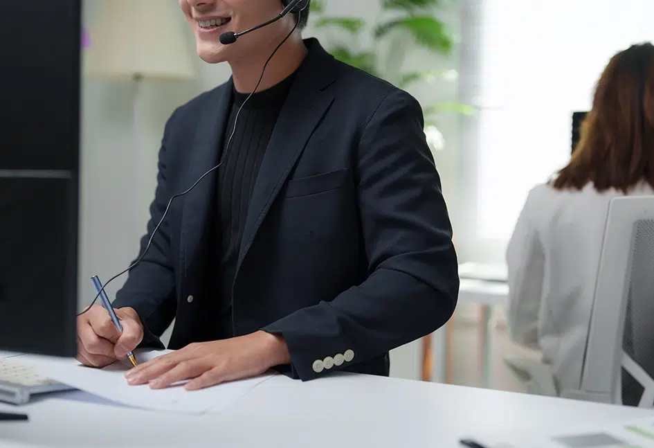 Lächelnder Mitarbeiter mit Headset beim Notizenschreiben während eines Kundensupport-Anrufs